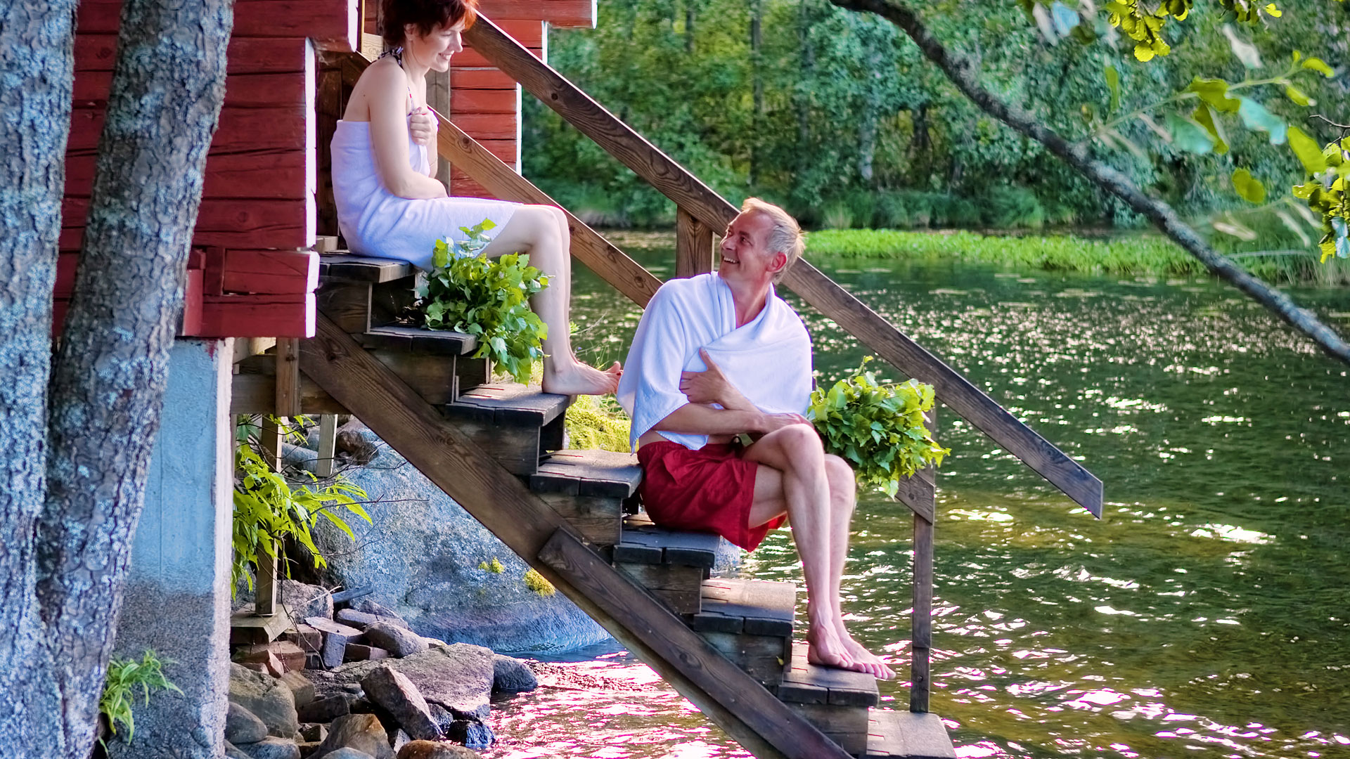 Mies ja nainen istuvat saunan rapuilla, jotka johtavat järveen. 