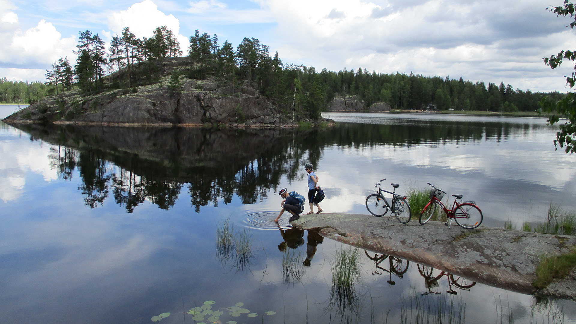 Kaksi pyöräilijää viettämässä taukoa matalalla rantakalliolla järven rannalla. 